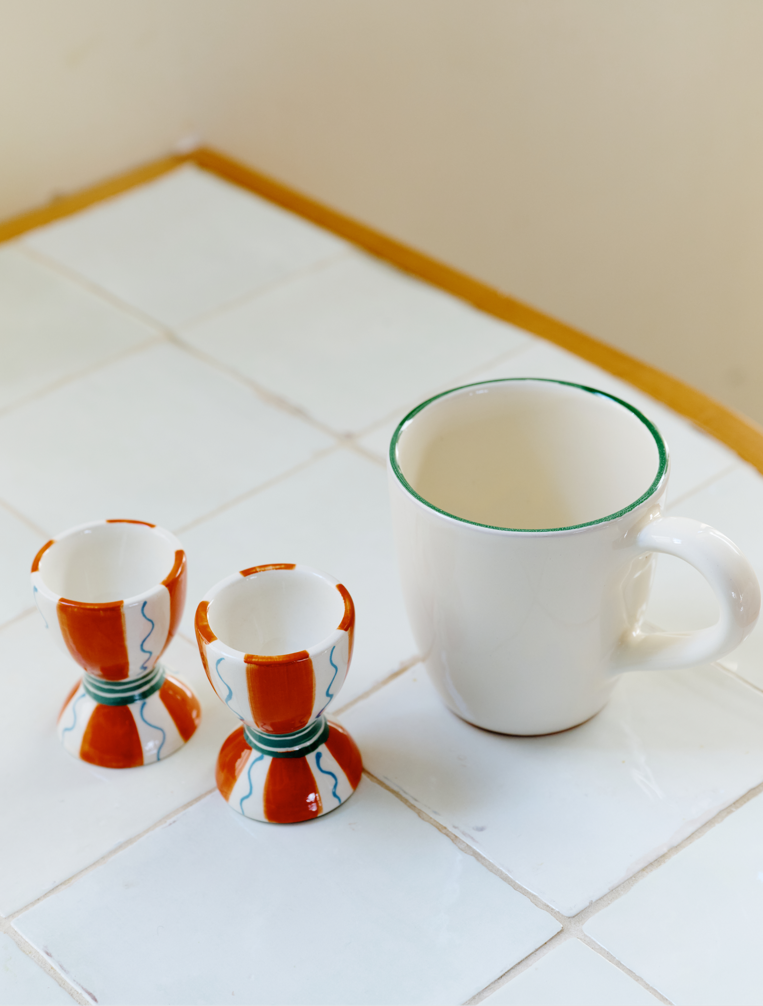 The Breakfast for Two Bundle features a white coffee mug with a green rim and two ceramic egg cups adorned with colorful red, blue, and green patterns—ideal for a cozy Valentine’s Day breakfast on a white tiled surface.
