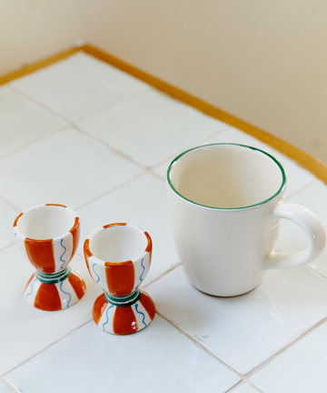 The Breakfast for Two Bundle features a white coffee mug with a green rim and two ceramic egg cups adorned with colorful red, blue, and green patterns—ideal for a cozy Valentine’s Day breakfast on a white tiled surface.