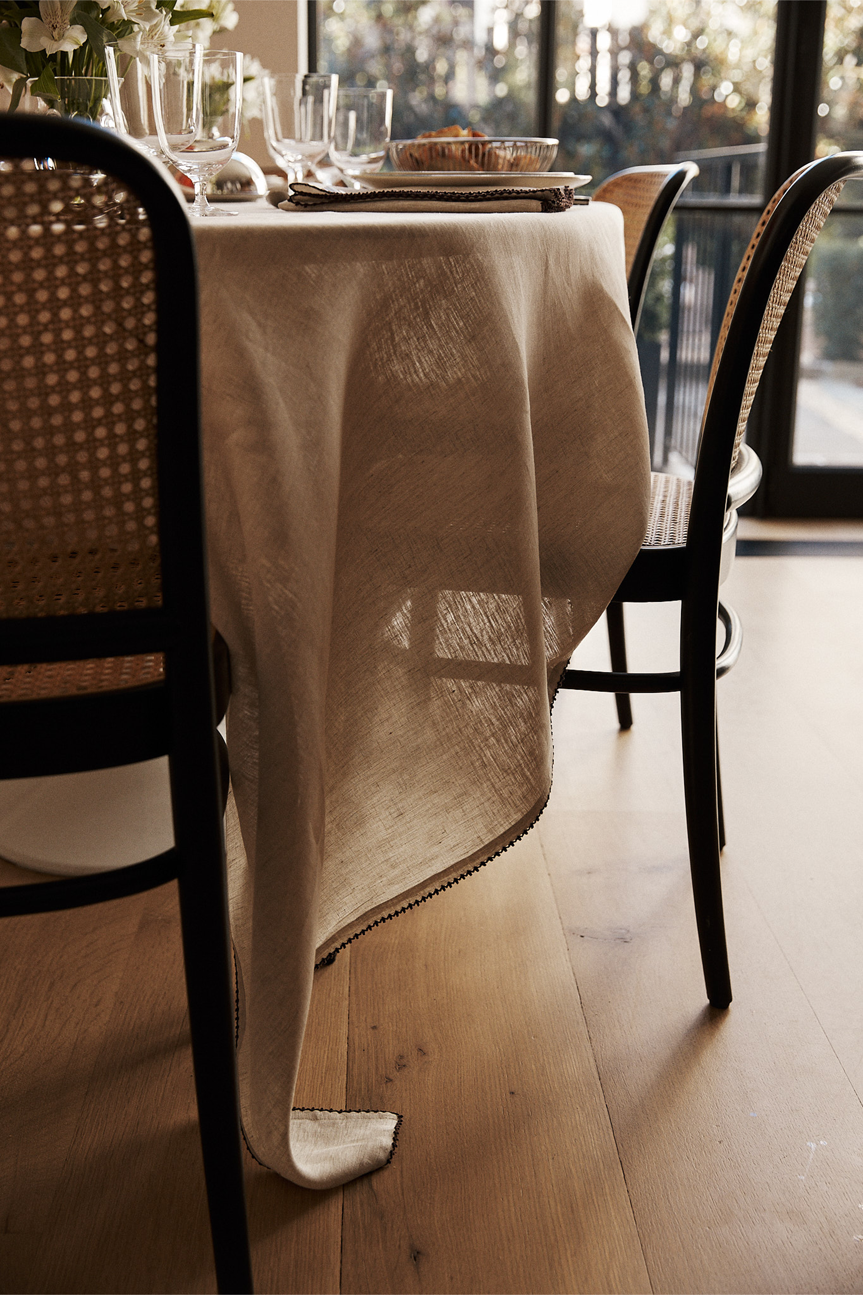 The Nico Tablecloth, hand-stitched linen, drapes a dining table set with plates and glasses, surrounded by wooden chairs with woven backs on a light wood floor, creating elegant table decor.