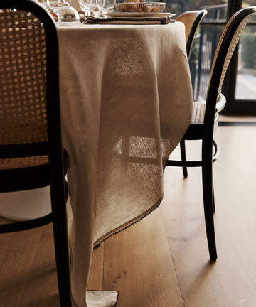 The Nico Tablecloth, hand-stitched linen, drapes a dining table set with plates and glasses, surrounded by wooden chairs with woven backs on a light wood floor, creating elegant table decor.