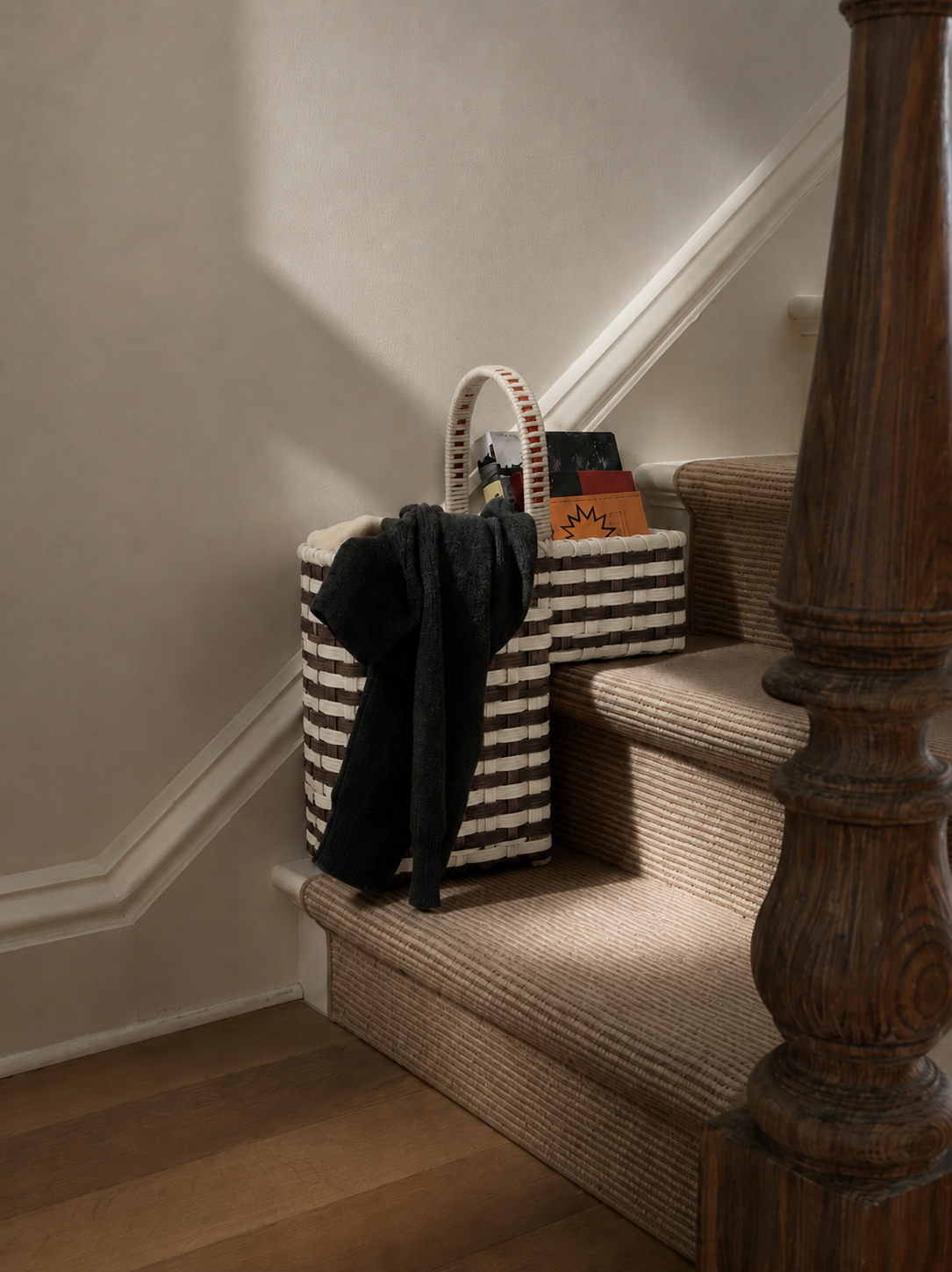 A Stair Tote featuring bold, handwoven stripes sits on a carpeted staircase, holding a black garment, a book, and assorted items—perfect for convenient storage on your stairs. Warm light casts soft shadows along the wall and wooden banister.
