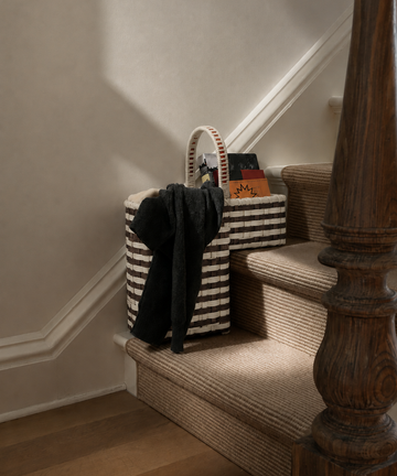 A Stair Tote featuring bold, handwoven stripes sits on a carpeted staircase, holding a black garment, a book, and assorted items—perfect for convenient storage on your stairs. Warm light casts soft shadows along the wall and wooden banister.