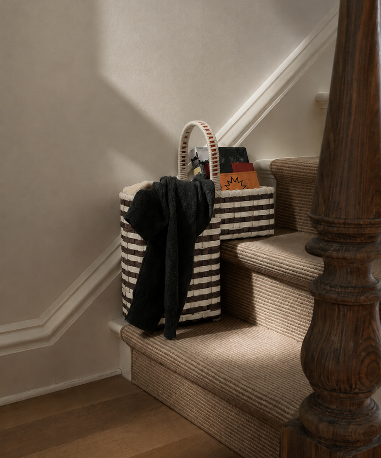 A Stair Tote featuring bold, handwoven stripes sits on a carpeted staircase, holding a black garment, a book, and assorted items—perfect for convenient storage on your stairs. Warm light casts soft shadows along the wall and wooden banister.