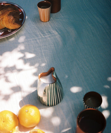 A white Filial Tablecloth with a delicate red branch stitch, topped with croissants, two brown cups, a striped milk jug, oranges—whole and peeled—and dappled sunlight across the scene.
