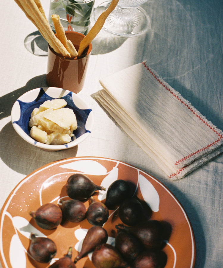 A tabletop scene features breadsticks in a cup, water glasses, neatly stacked Filial Napkin, Set of 6 cotton napkins, a bowl of cheese, and fresh figs on a patterned plate.