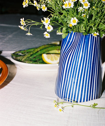 The Natalia Water Jug, featuring blue and white stripes with small white flowers, is displayed on a table beside a plate of asparagus and lemon slices.