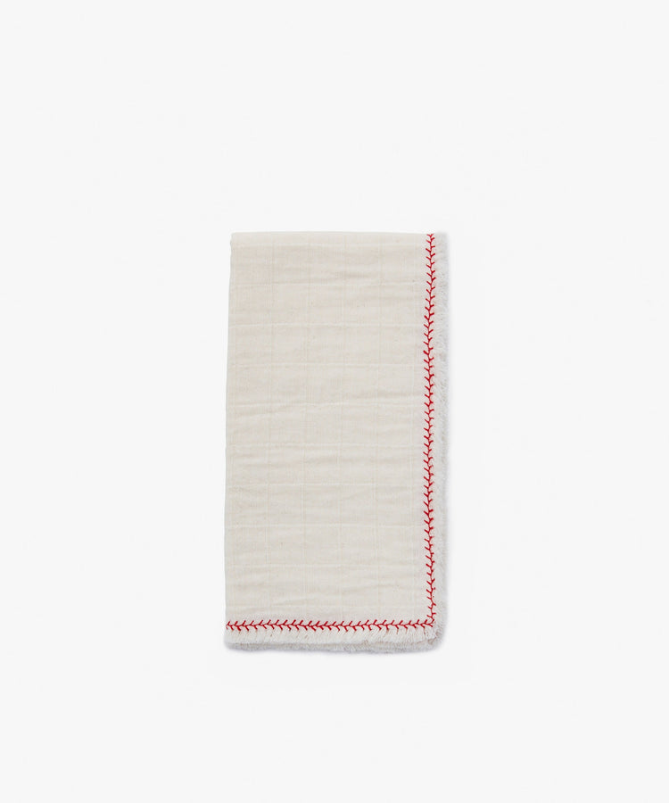 A folded Filial Napkin, Set of 6, in off-white cotton with a red decorative edge stitch, rests on a plain white tabletop.